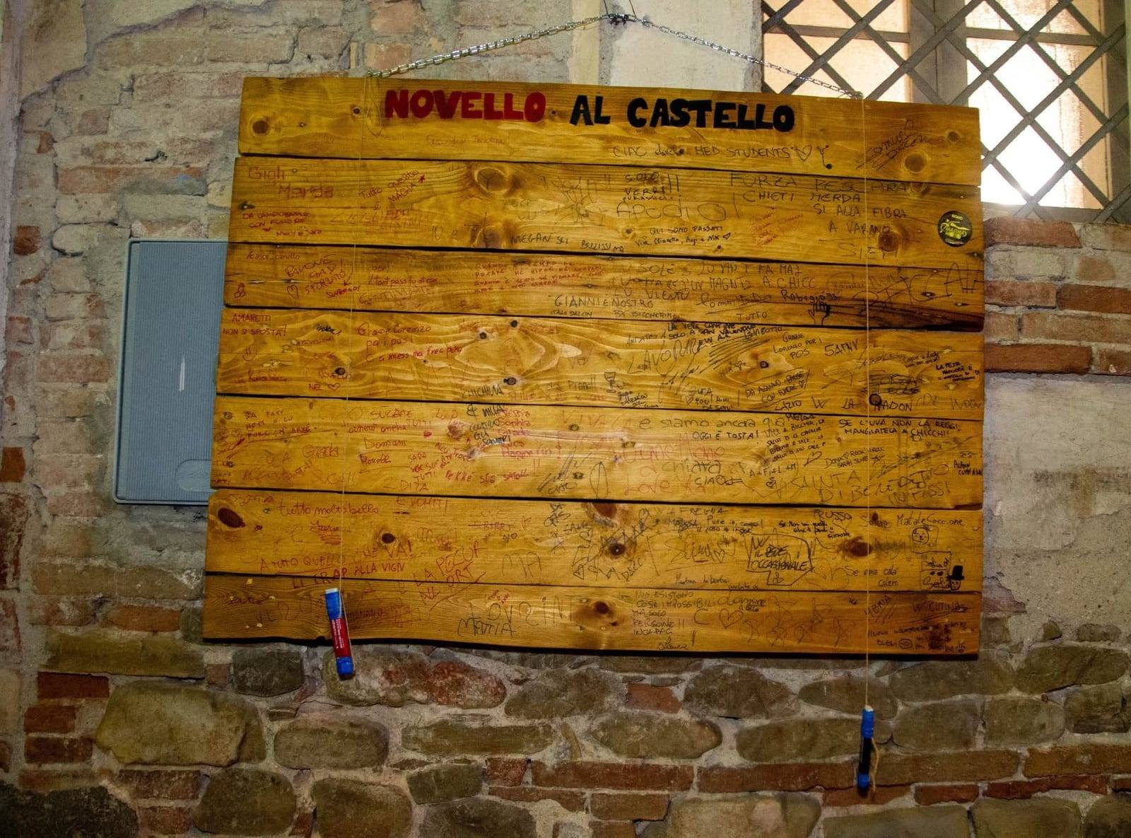 Pannello in legno con la scritta “Novello al Castello” su un muro in pietra