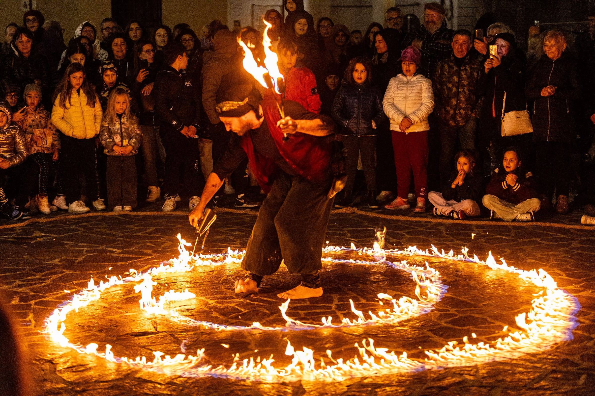 Un’artista danza al centro di una spirale di fuoco, circondata dal pubblico in piazza