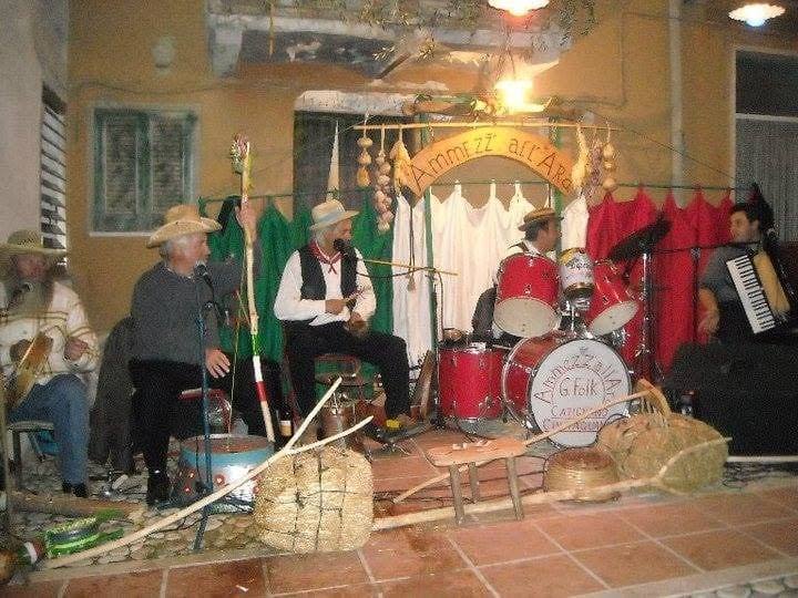 Un gruppo folk con cappelli di paglia, batteria e fisarmonica su un palco decorato con paglia e tende verdi e rosse